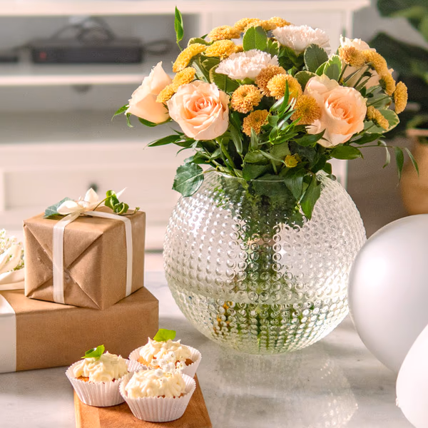A bunch of flowers in a vase, next to presents and cupcakes