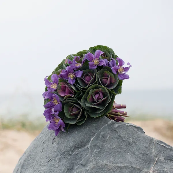 purple and green bouquet lies upon a rock at the beach with sand and water in the background
