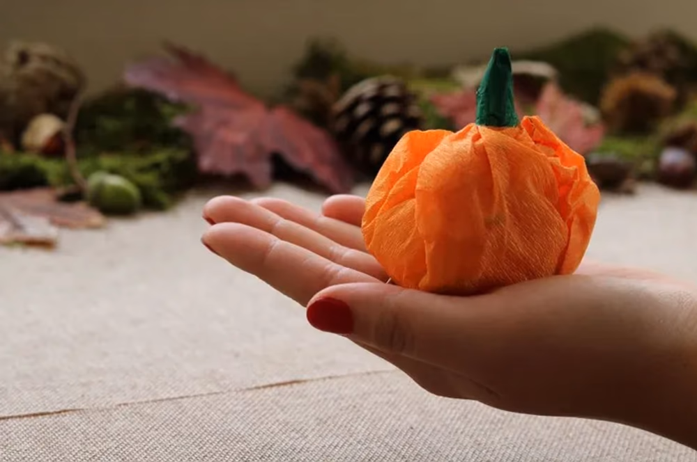 A hand holding the completed paper pumpkin decoration with autumn leaves and pine cones in the background
